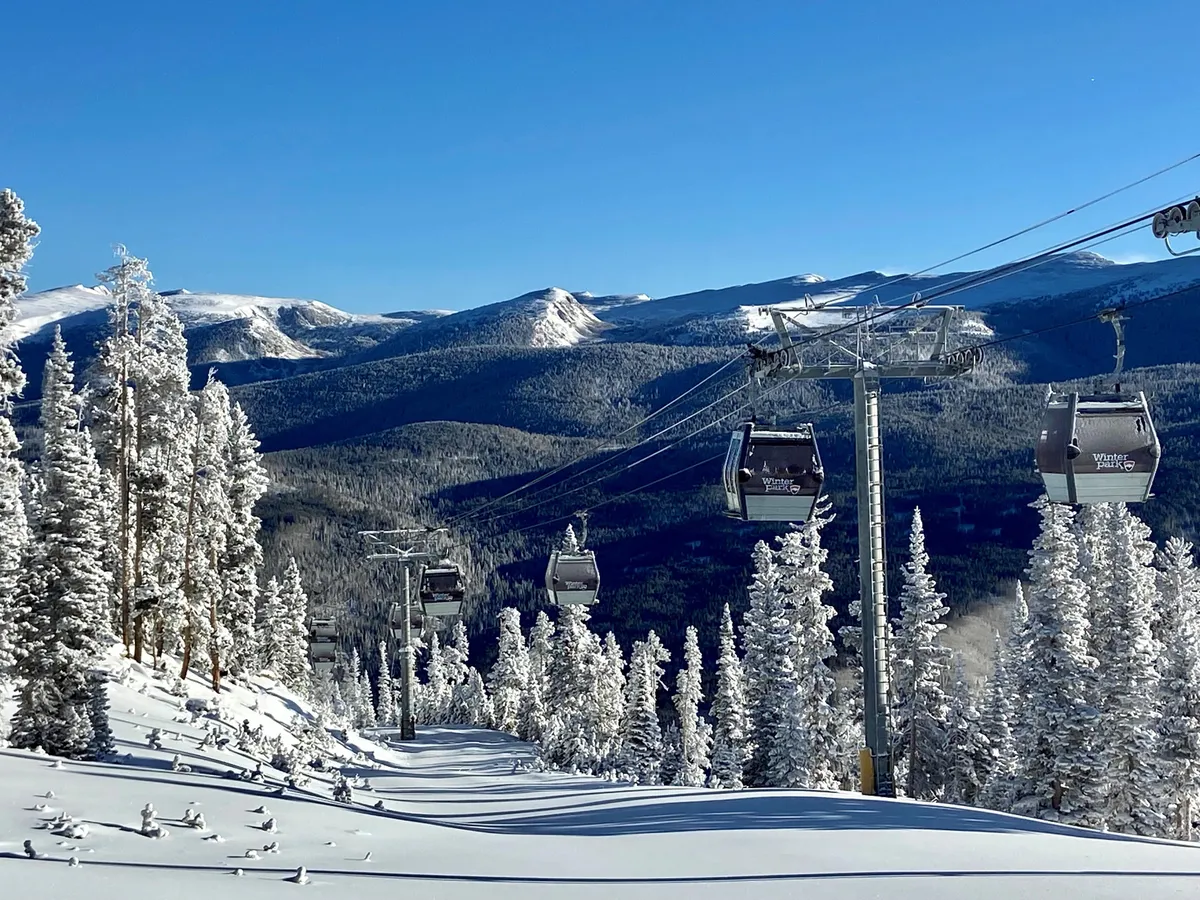 A breathtaking exterior view of snow-covered mountains with a prominent gondola lift gliding across the scene. Pine trees dusted with snow frame the landscape, while a wide expanse of blue sky suggests a crisp, clear winter day. The image evokes a sense of adventure and the allure of world-class skiing and snowboarding right outside your door.