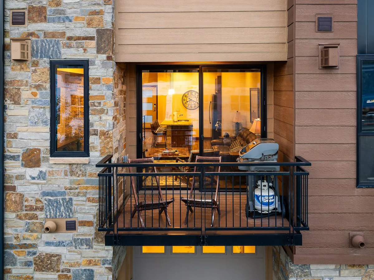 A cozy balcony setup with comfortable seating and a grill, illuminated by warm interior lights. The space invites guests to relax outdoors, dine al fresco, or share stories as the sun sets.