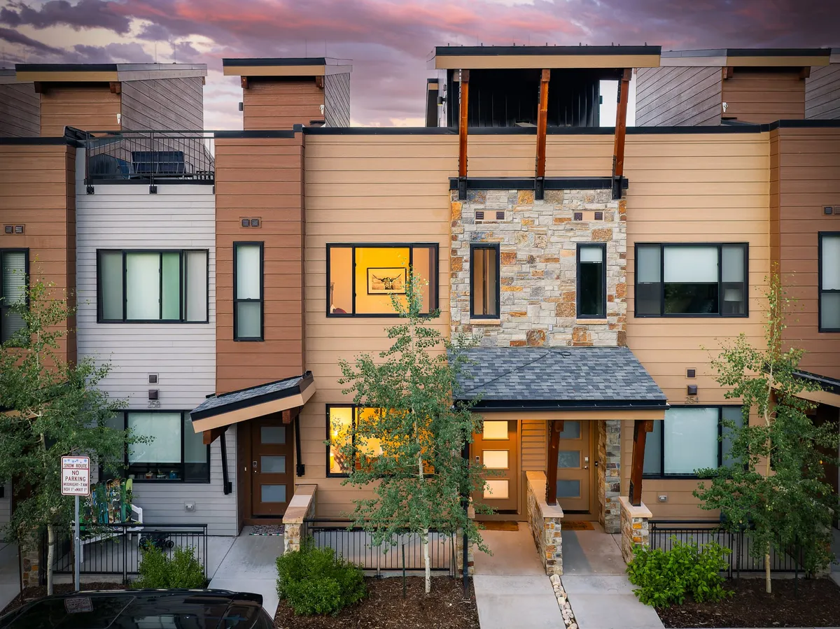 The image showcases the stunning exterior of a luxury single-family home, featuring a striking stone façade, rustic wooden beams, expansive windows, and a beautifully landscaped front yard. The home is bathed in the warm glow of sunset, hinting at breathtaking mountain views from the balcony and inviting spaces for evening gatherings or family barbecues.