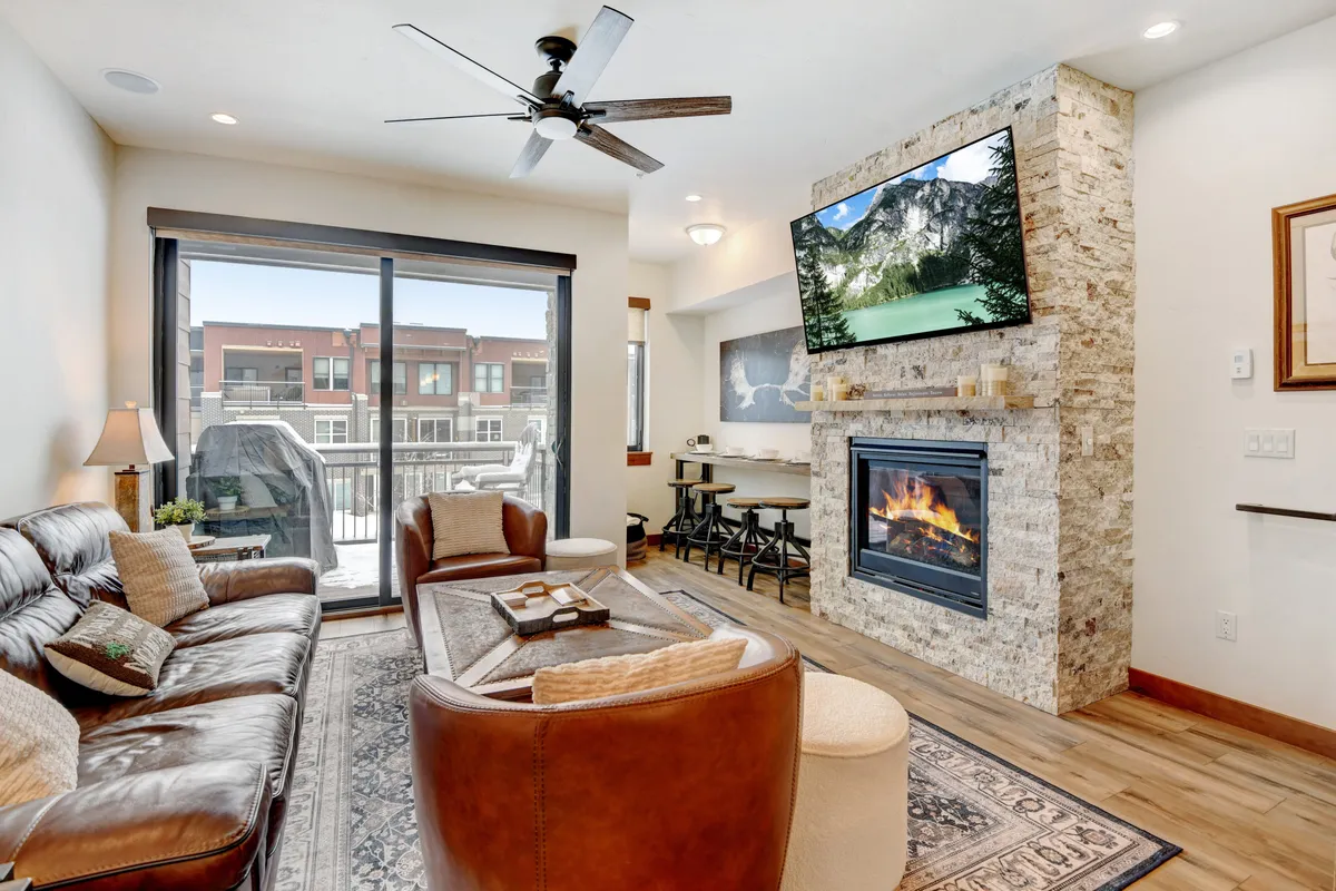 A warm, inviting open-plan living space featuring a comfortable leather sofa positioned by a striking stone fireplace, with a large TV set above. The dining area with tasteful artwork is visible just beyond, creating a seamless flow between relaxation, entertainment, and communal meals. Expansive windows allow natural mountain light to stream in, enhancing the cozy atmosphere.