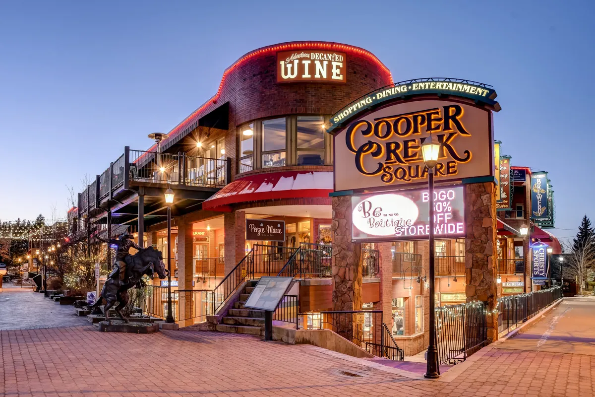 A lively exterior of Cooper Creek Square, featuring a prominent sign, a wine shop, inviting outdoor seating, and festive decorative lighting. The area bustles with the charm of a mountain town, hinting at shopping, dining, and vibrant community events.