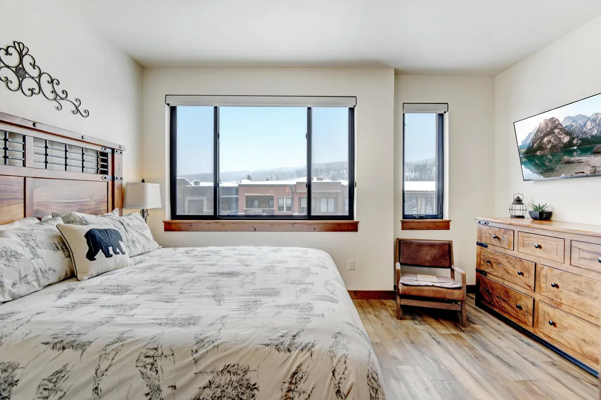 A tranquil bedroom scene featuring a large bed with patterned bedding, expansive windows offering a mountain view, a wooden dresser, and a comfortable chair by the window. The space exudes calmness and warmth, ideal for private rest and relaxation.