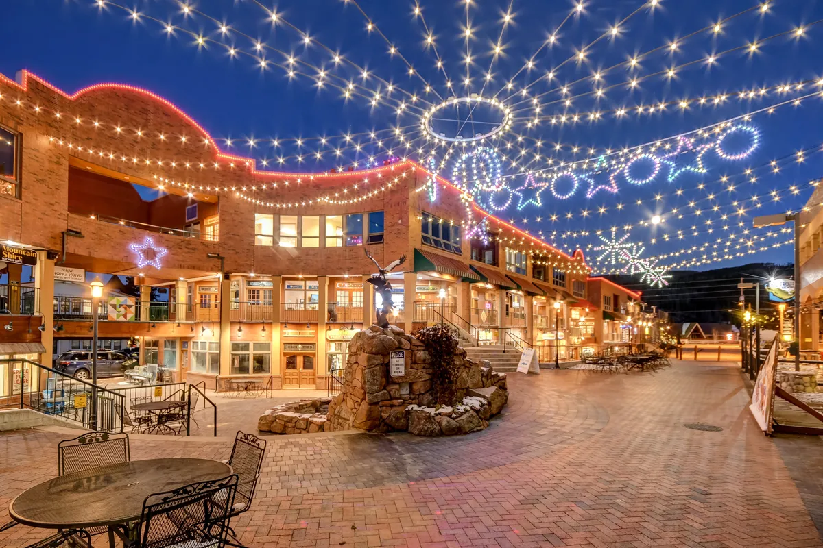 A lively downtown scene featuring decorative string lights, a brick pathway, outdoor seating, a stone fountain, and inviting storefronts. The area looks bustling and festive, perfect for dining al fresco, shopping, and enjoying social gatherings.